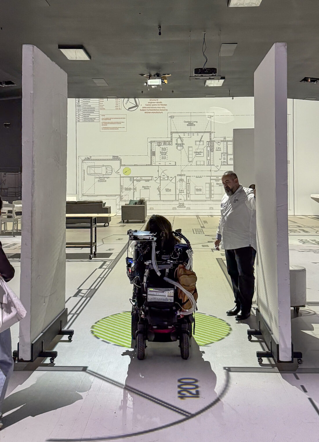 A participant drives a power wheelchair through a full-scale doorway opening while a designer looks on, with clearance dimensions and a turning circle projected on the floor at Lifesize Plans Burwood